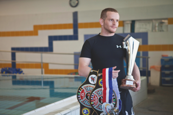 Itai Liptz, a multi-time world champion, stands beside an indoor pool holding a championship trophy, medals, and title belts while discussing the value of mentorship.