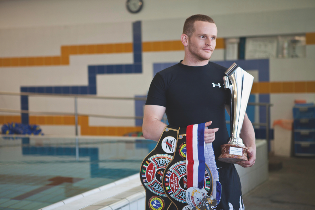 Itai Liptz, a multi-time world champion, stands beside an indoor pool holding a championship trophy, medals, and title belts while discussing the value of mentorship.