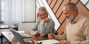 Two older adults, a woman and a man, sit together at a desk smiling while working on papers and using laptops.