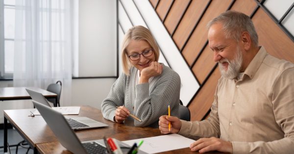 Two older adults, a woman and a man, sit together at a desk smiling while working on papers and using laptops.